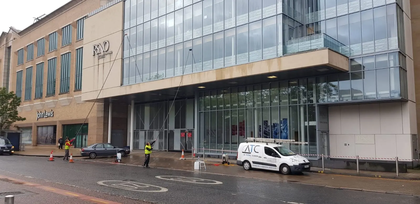 ATC team cleaning a glazed city-centre frontage from the pavement with water-fed poles, the van parked alongside behind a cordon