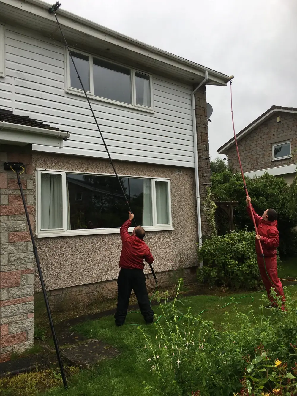 Two cleaners in red jackets washing fascias and soffits on a semi-detached house